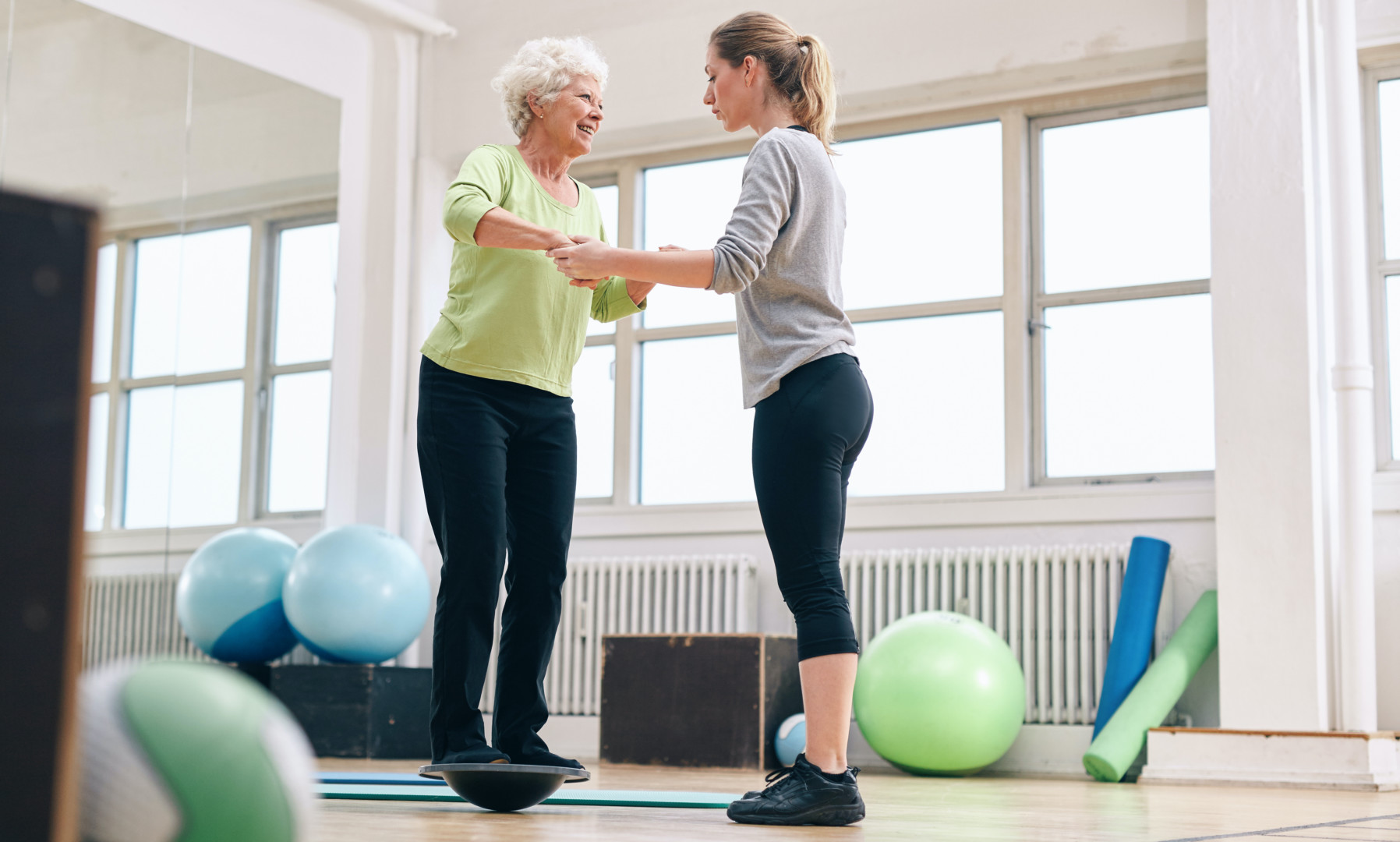 Female trainer helping senior woman in a gym exercising with a bosu balance training platform. Elder woman being assisted by gym instructor while workout session.