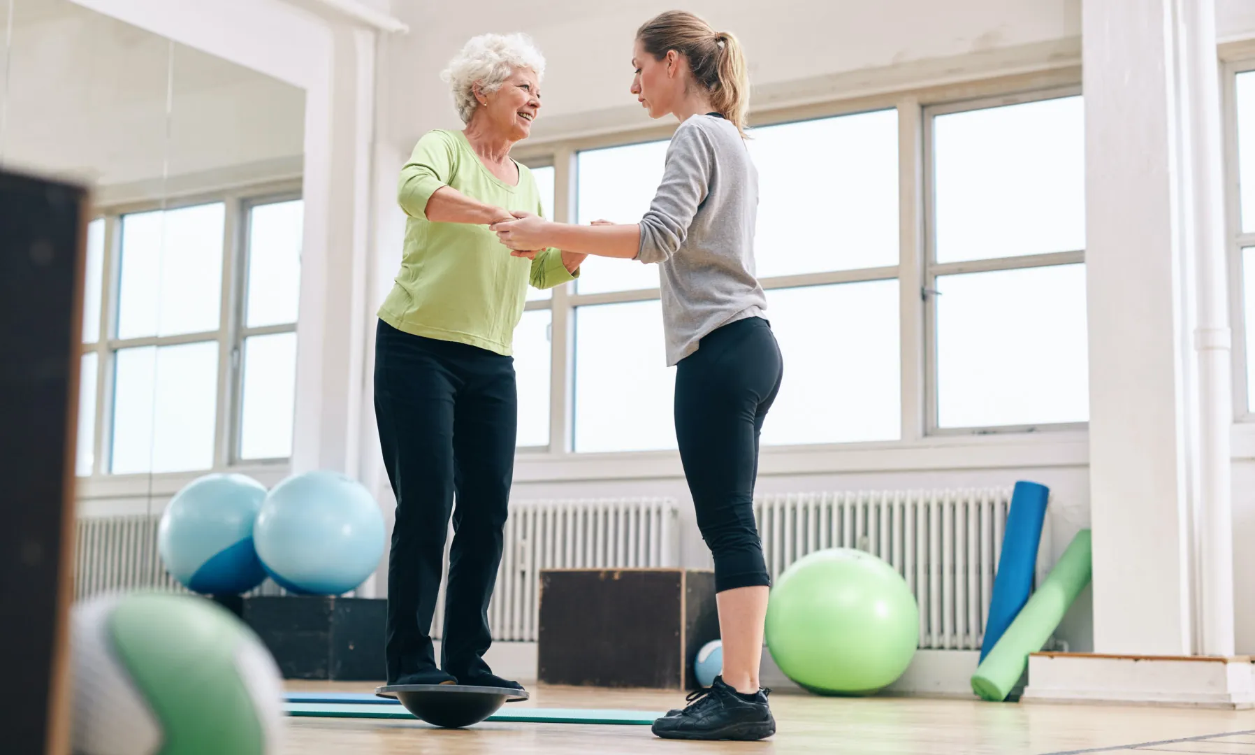 Trainer helping senior woman on bosu balance training platform Female trainer helping senior woman in a gym exercising with a bosu balance training platform. Elder woman being assisted by gym instructor while workout session.